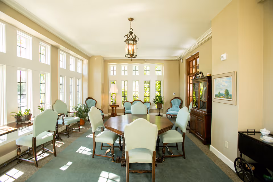 A bright and elegant sitting room with large windows letting in natural light. The room features a wooden octagonal table surrounded by upholstered chairs with light blue fabric. Additional matching armchairs are placed along the walls. There are several potted plants, a floor lamp, a wooden cabinet with glass doors, and a framed painting on the wall. The room has beige walls and a green carpet.