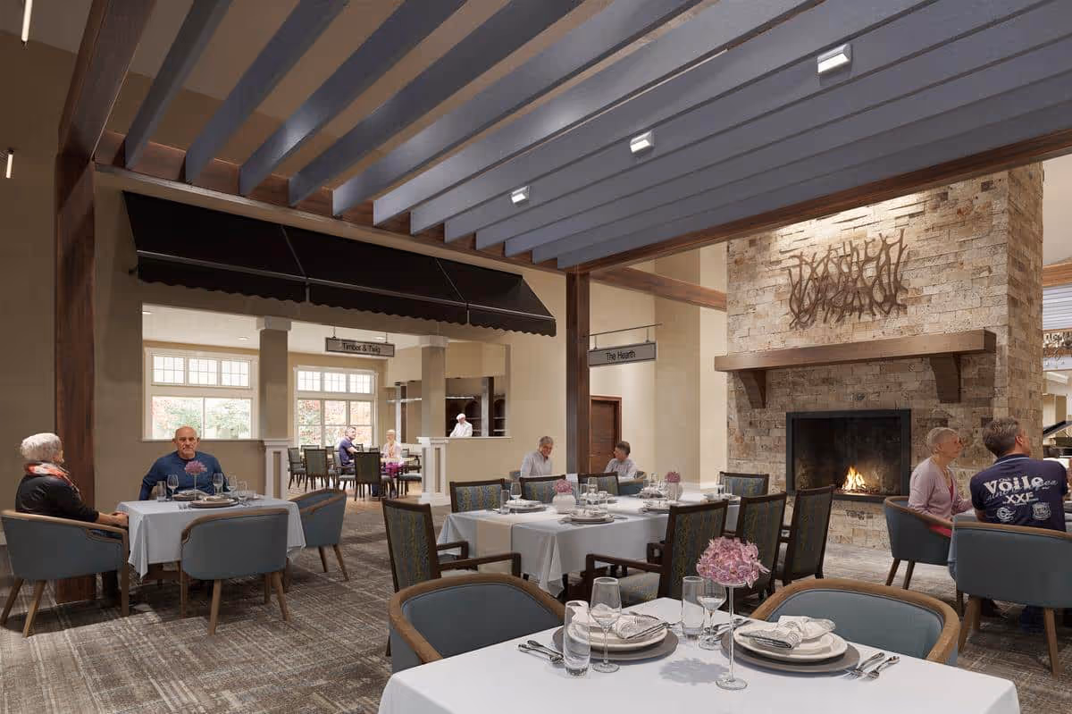 A cozy dining room in The Timberley facility with several tables set with white tablecloths, plates, glasses, and silverware. There are elderly people seated at some tables, engaging in conversation. The room features a large stone fireplace with a fire burning, wooden beams on the ceiling, and soft lighting creating a warm atmosphere.