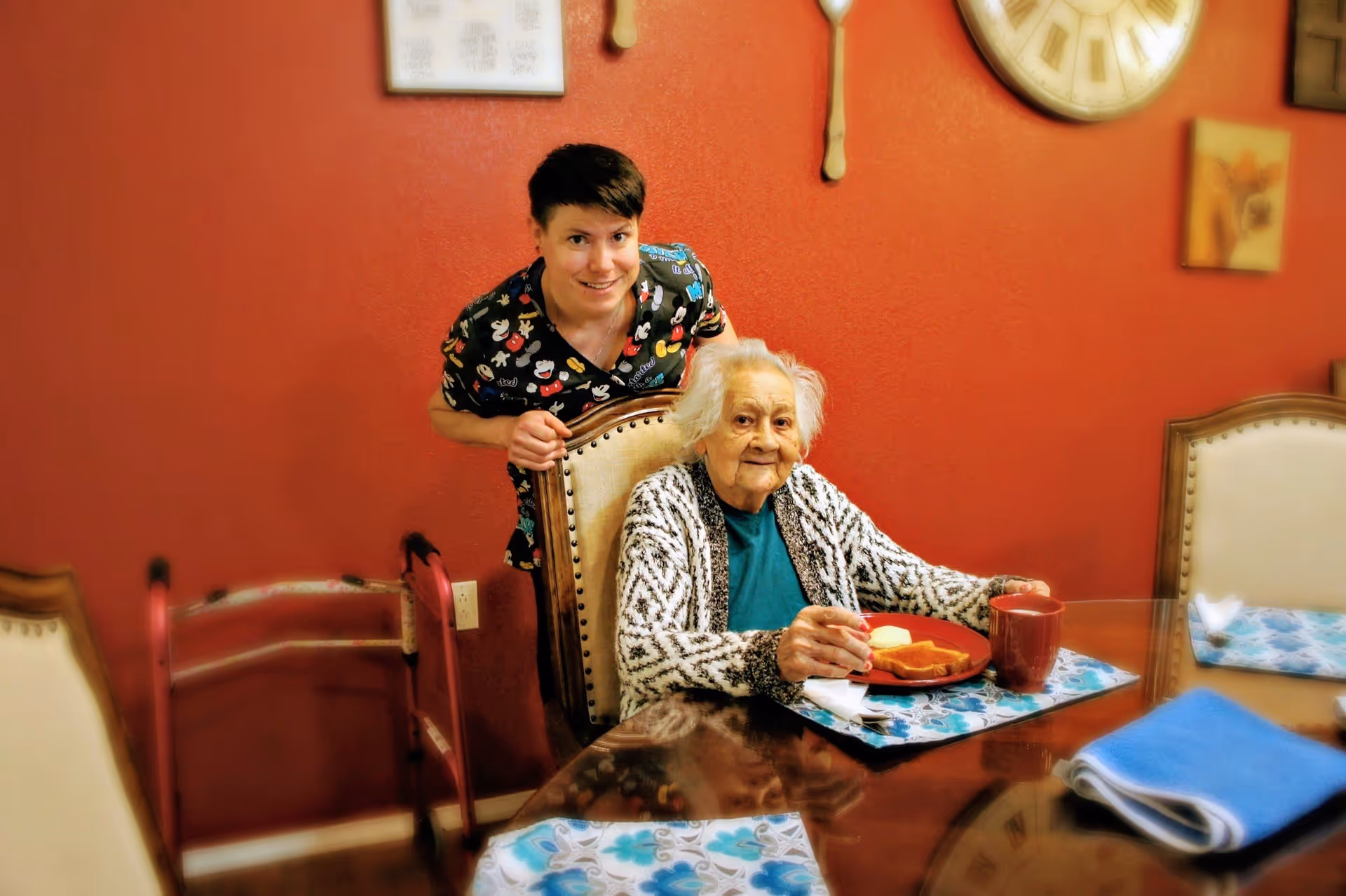 An elderly woman seated at a dining table with a caregiver standing behind her, a plate and mug on the table.