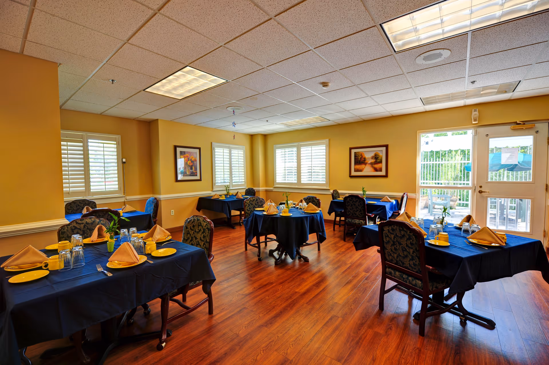 A dining room with several tables covered in dark blue tablecloths, each set with yellow plates, folded napkins, glasses, and silverware. The room has wooden flooring, yellow walls with framed artwork, and windows with white shutters. A glass door leads outside to a patio area.