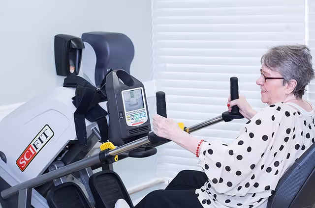An elderly woman with short gray hair and glasses is using a SciFit exercise machine in a bright room with white blinds in the background. She is seated and holding the machine's handles, engaging in a workout.