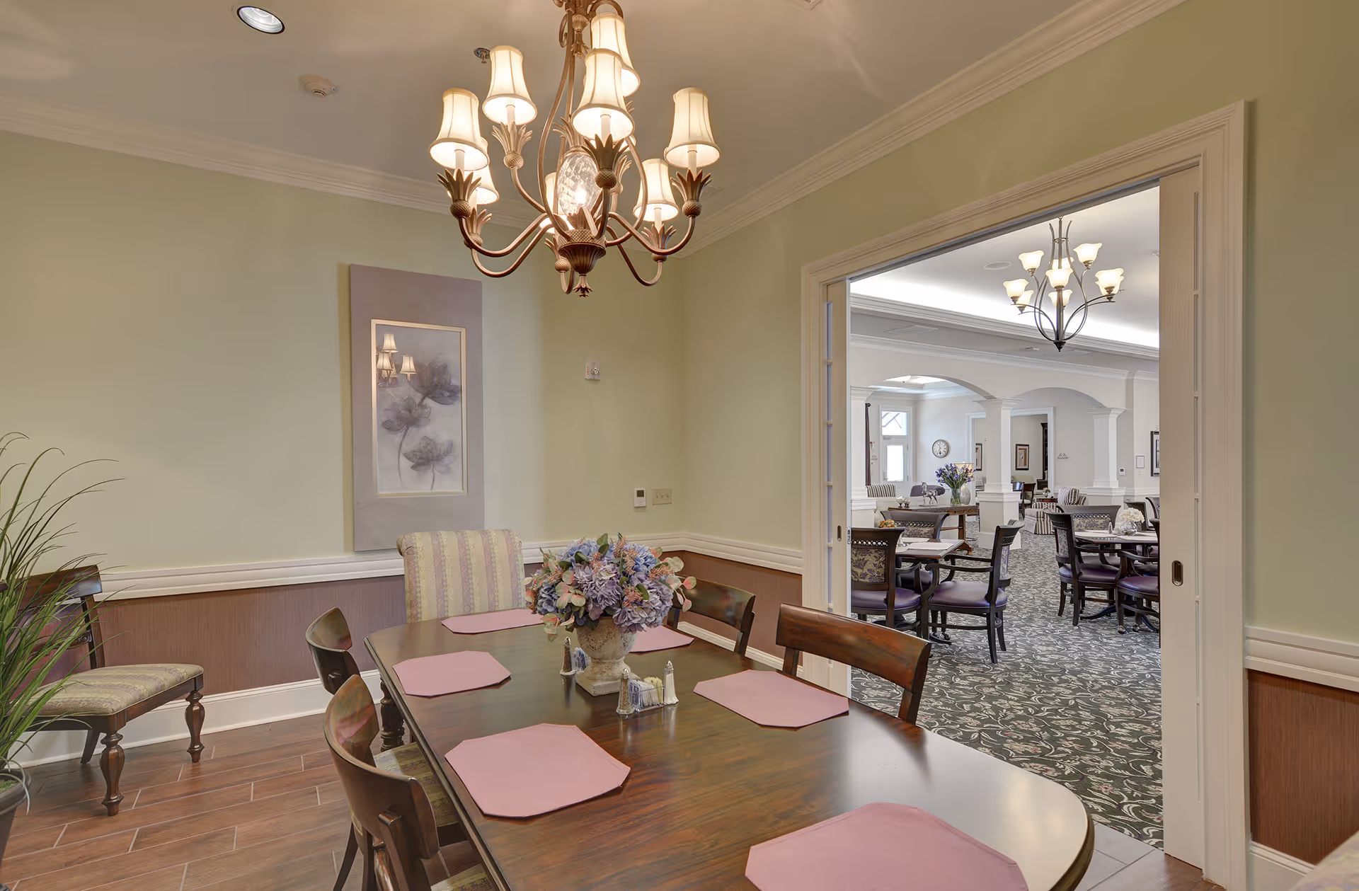 A dining area with a wooden table set with six pink placemats and a floral centerpiece. The room has light green walls, a chandelier with multiple lampshades, and a framed artwork on the wall. Through an open doorway, another larger dining or common area with multiple tables and chairs is visible, featuring patterned carpet and another chandelier.