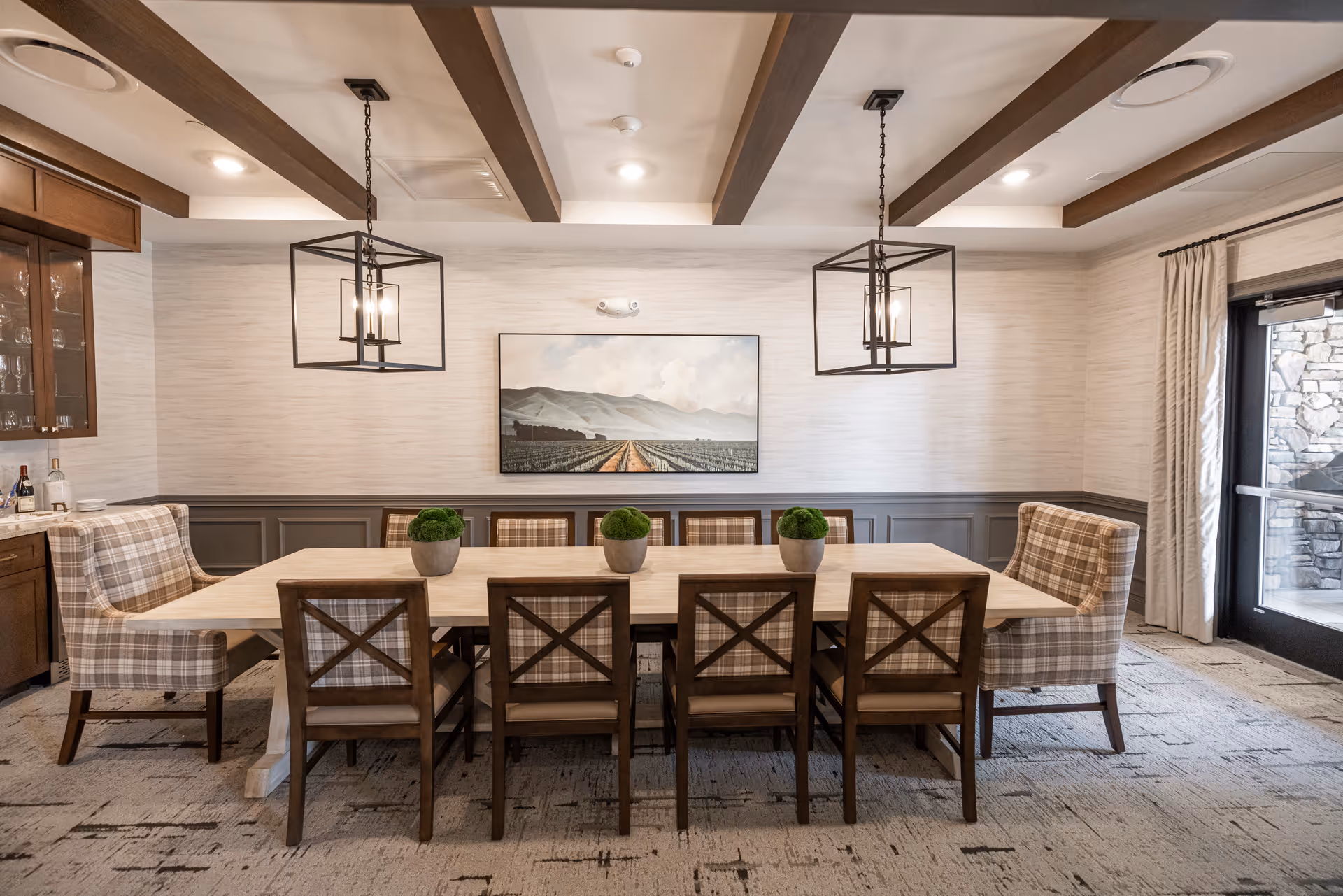 Dining room with a long wooden table surrounded by wooden chairs and plaid upholstered armchairs, two pendant light fixtures, potted plants, and a landscape painting on the wall.