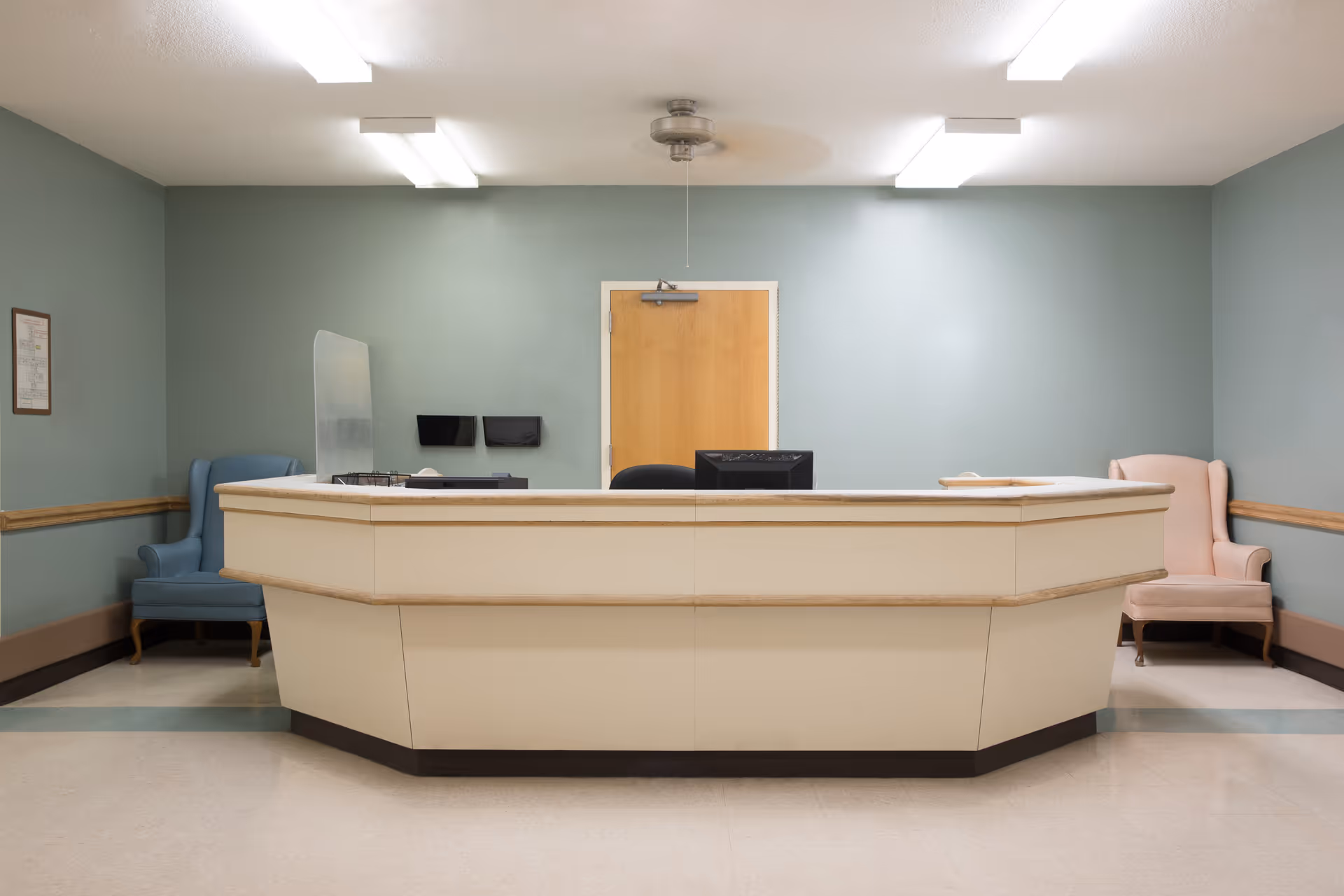 A beige reception desk in a small lobby with two upholstered armchairs against mint-green walls and a closed wooden door behind it.