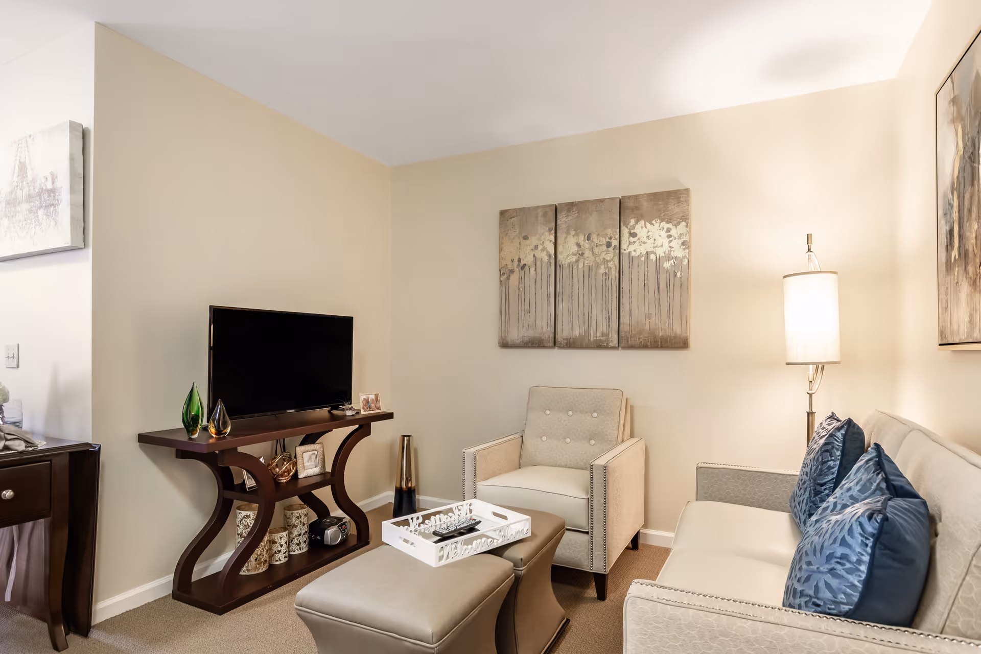 A cozy living room with a beige armchair and sofa adorned with blue patterned pillows. A modern wooden TV stand holds a flat-screen television and decorative items. A white tray with remote controls sits on a beige ottoman. The walls are light beige with three-panel artwork above the armchair and a floor lamp providing warm lighting.