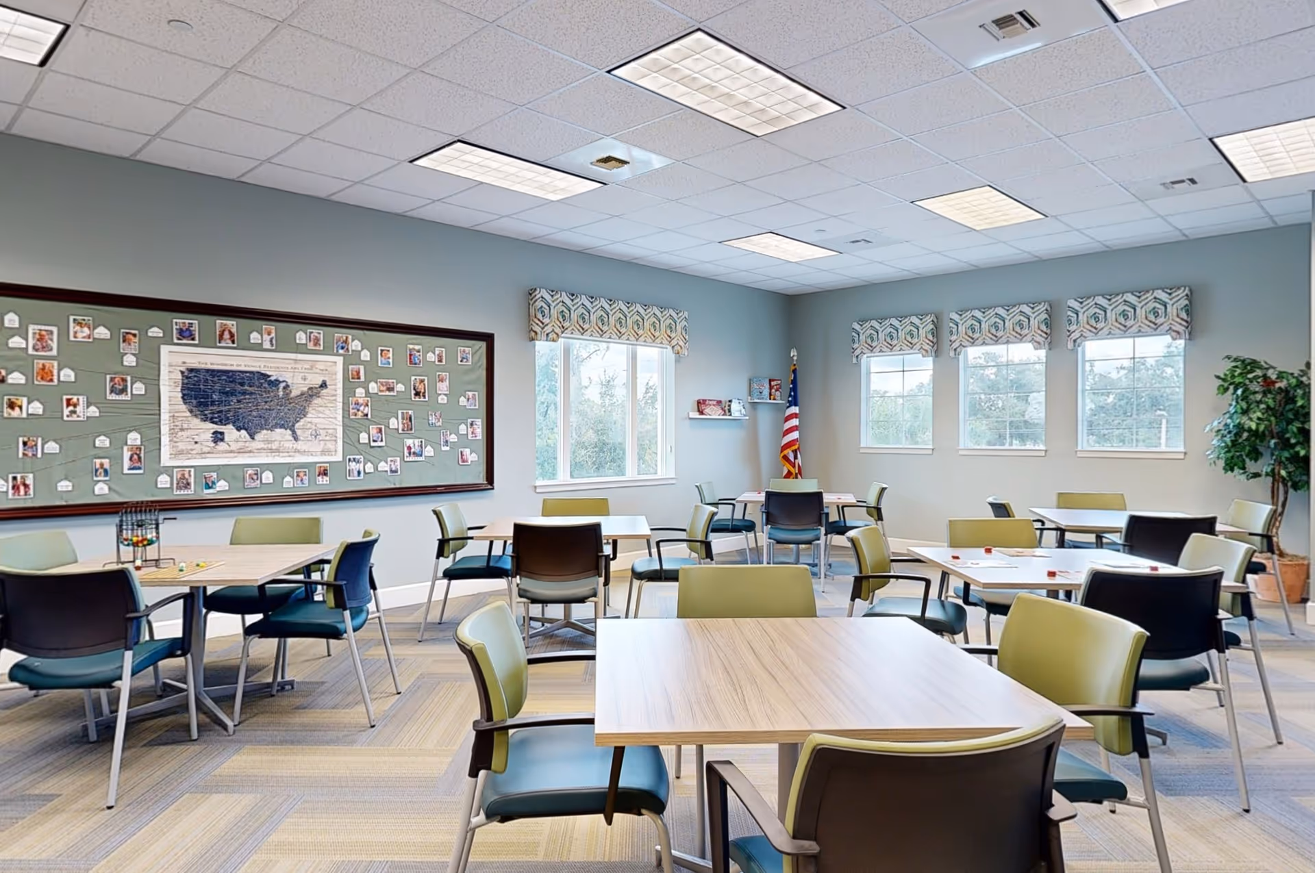 A bright and spacious activity or common room with multiple tables and chairs arranged for group seating. The room has large windows with patterned valances, a bulletin board with photos and a map of the United States, an American flag in the corner, and a potted plant. The ceiling has recessed lighting and a tiled design.