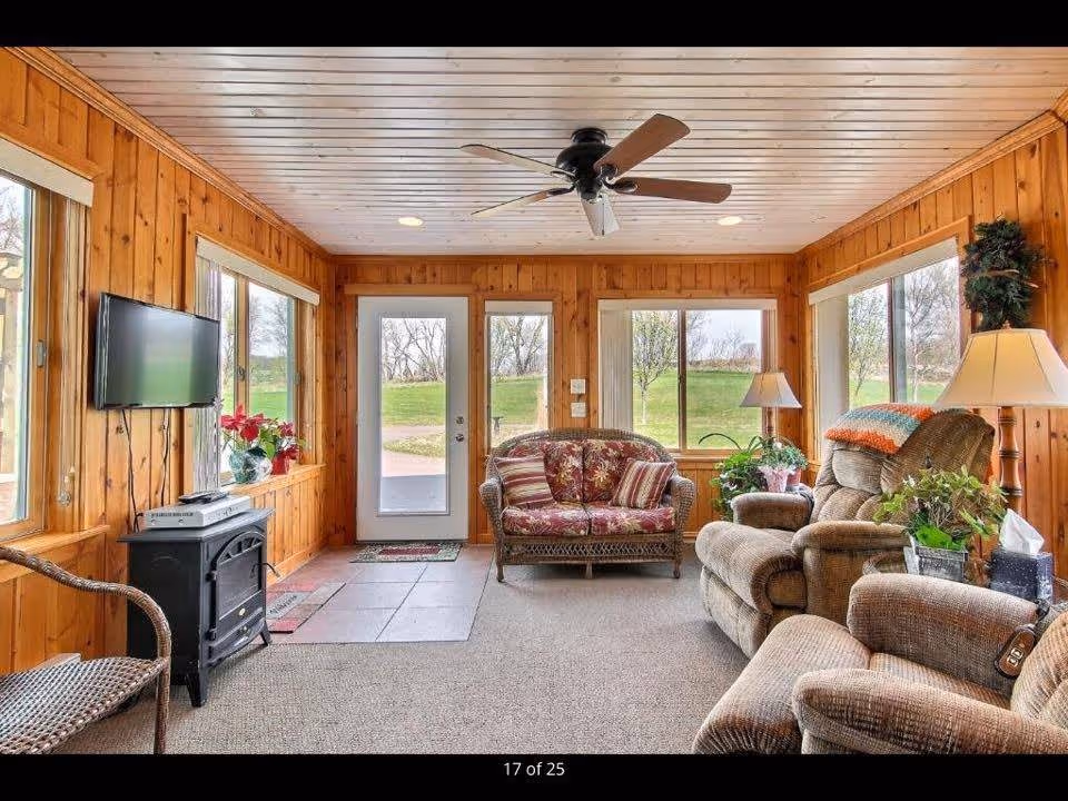 A cozy living room with wood-paneled walls and ceiling, featuring a ceiling fan, large windows with views of green outdoor space, a glass door, a wall-mounted flat-screen TV above a small black electric fireplace, a wicker loveseat with floral cushions, two recliner chairs, a side table with a lamp, and several potted plants.