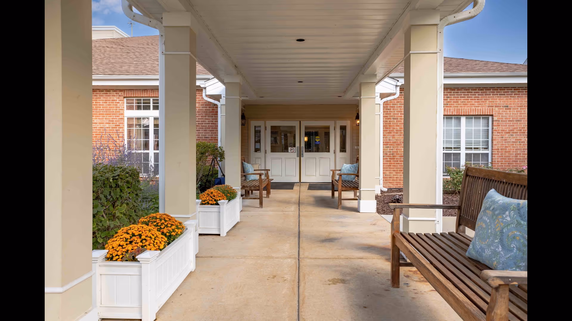 Covered entrance to a brick senior living building with benches, white planters of orange flowers, and double doors.