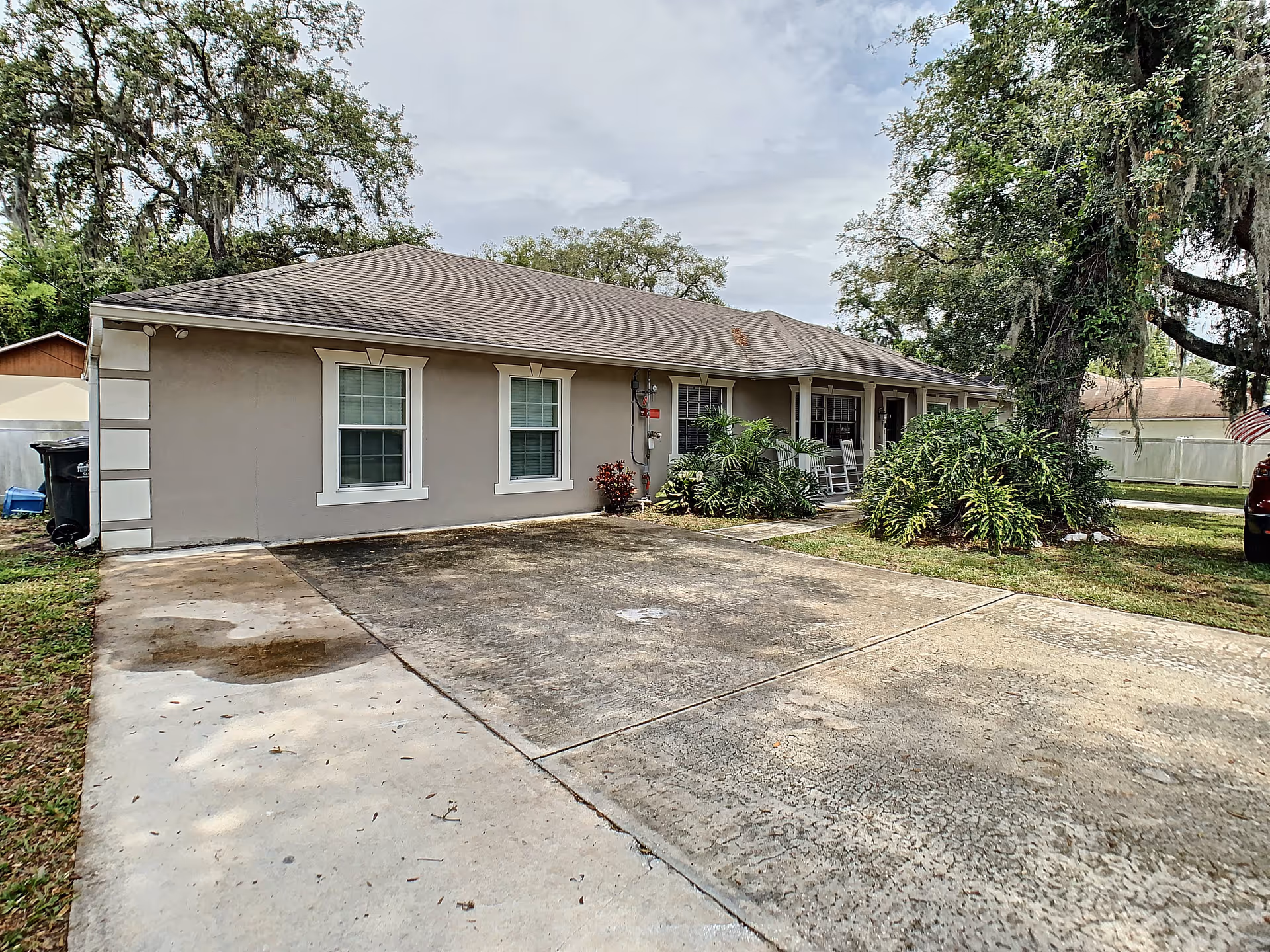 Single-story residential building with a gray exterior, white trim around windows, and a shingled roof. There is a concrete driveway in front, some greenery including bushes and a large tree with hanging moss, and a covered porch with rocking chairs.