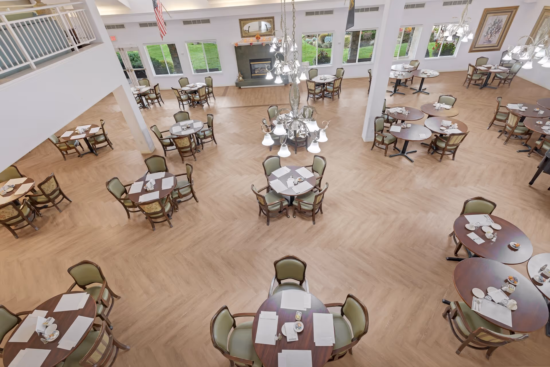 A spacious dining room with multiple round wooden tables, each surrounded by four chairs with green cushions. The tables are set with white napkins, cups, and small plates. The room has large windows letting in natural light, a fireplace with a mirror above it, and two chandeliers hanging from the ceiling. The floor is light wood with a herringbone pattern.