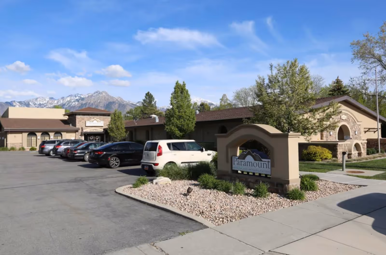 Exterior view of Paramount Health and Rehabilitation facility with a parking lot containing several cars. The building is surrounded by trees and landscaping with mountains visible in the background under a blue sky with some clouds.