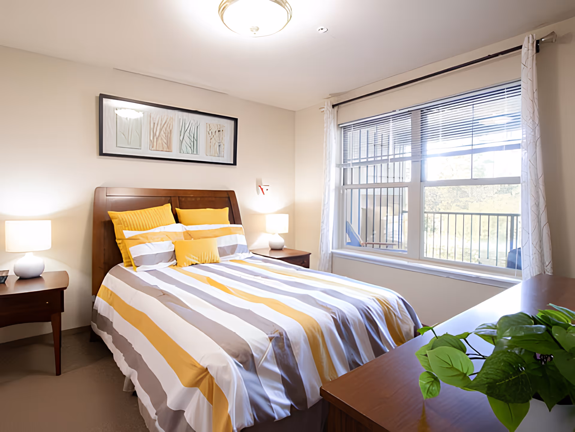 A bright bedroom with a double bed featuring yellow, white, and gray striped bedding and multiple pillows. There are two bedside tables with lamps on either side of the bed. A framed artwork hangs above the headboard. A large window with blinds and white curtains lets in natural light, and a green potted plant sits on a wooden dresser in the foreground.