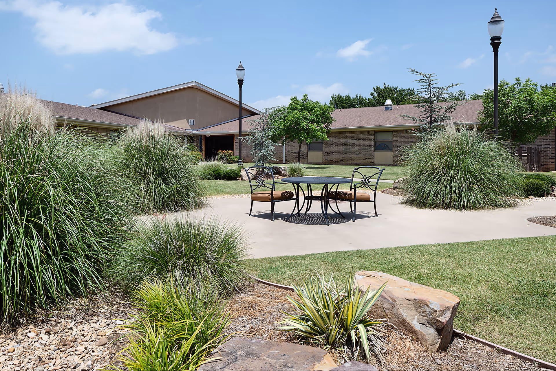 Sunlit courtyard with a round metal table and chairs surrounded by ornamental grasses and single-story brick buildings.