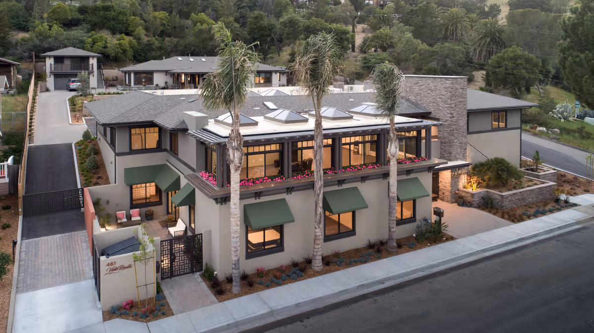 Two-story modern senior living building with palm trees, green window awnings, a flower-lined balcony, and a gated landscaped entrance.