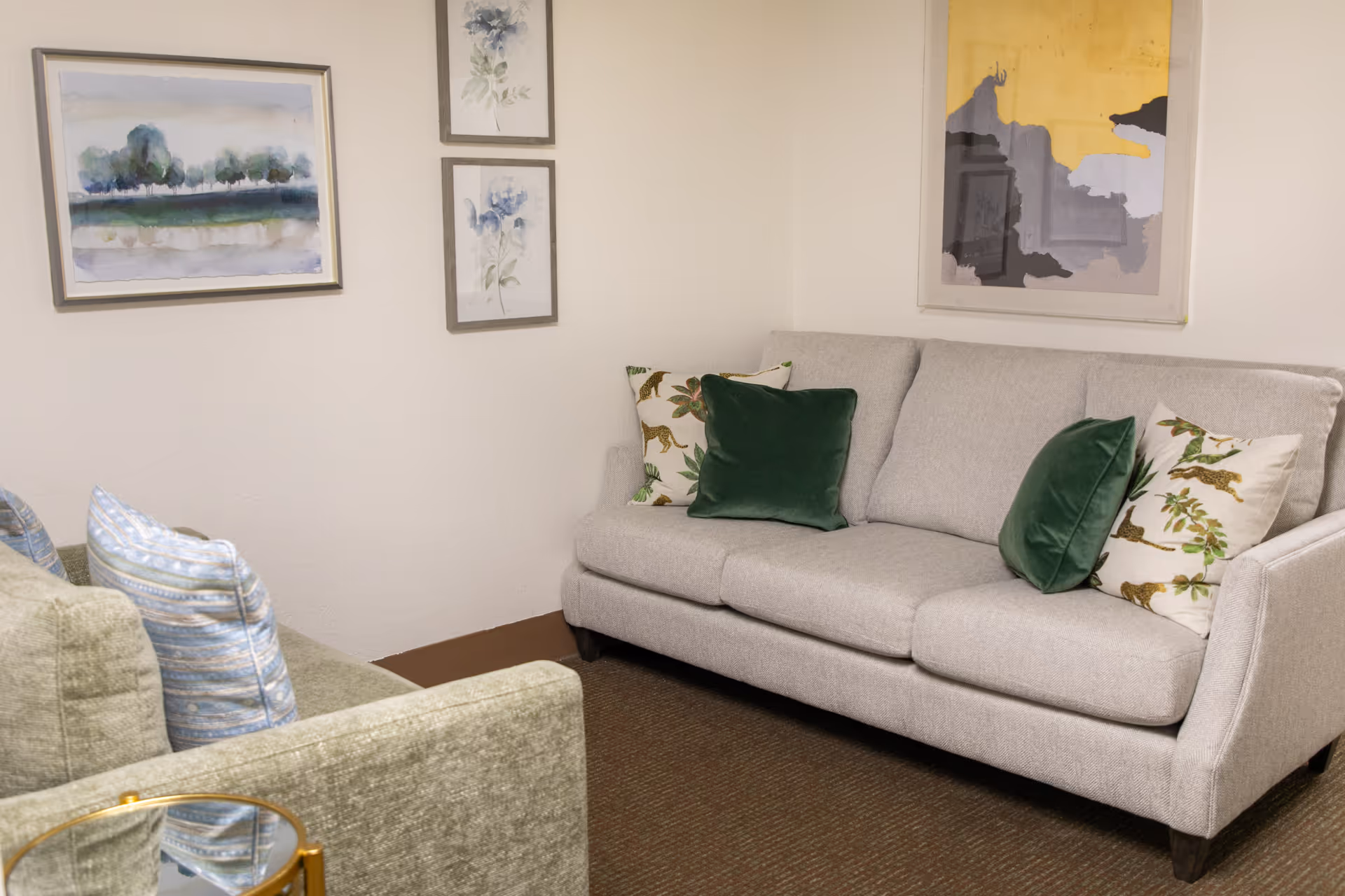 A small living room with a gray sofa and green throw pillows, a second upholstered chair partially visible, and framed artwork on the walls.