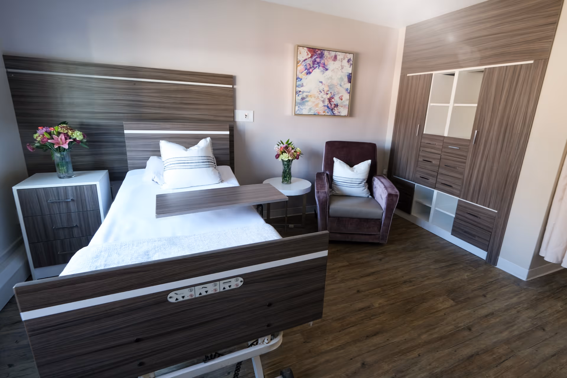A furnished senior living bedroom with a hospital-style bed, nightstand, armchair, flowers, and a built-in wardrobe.