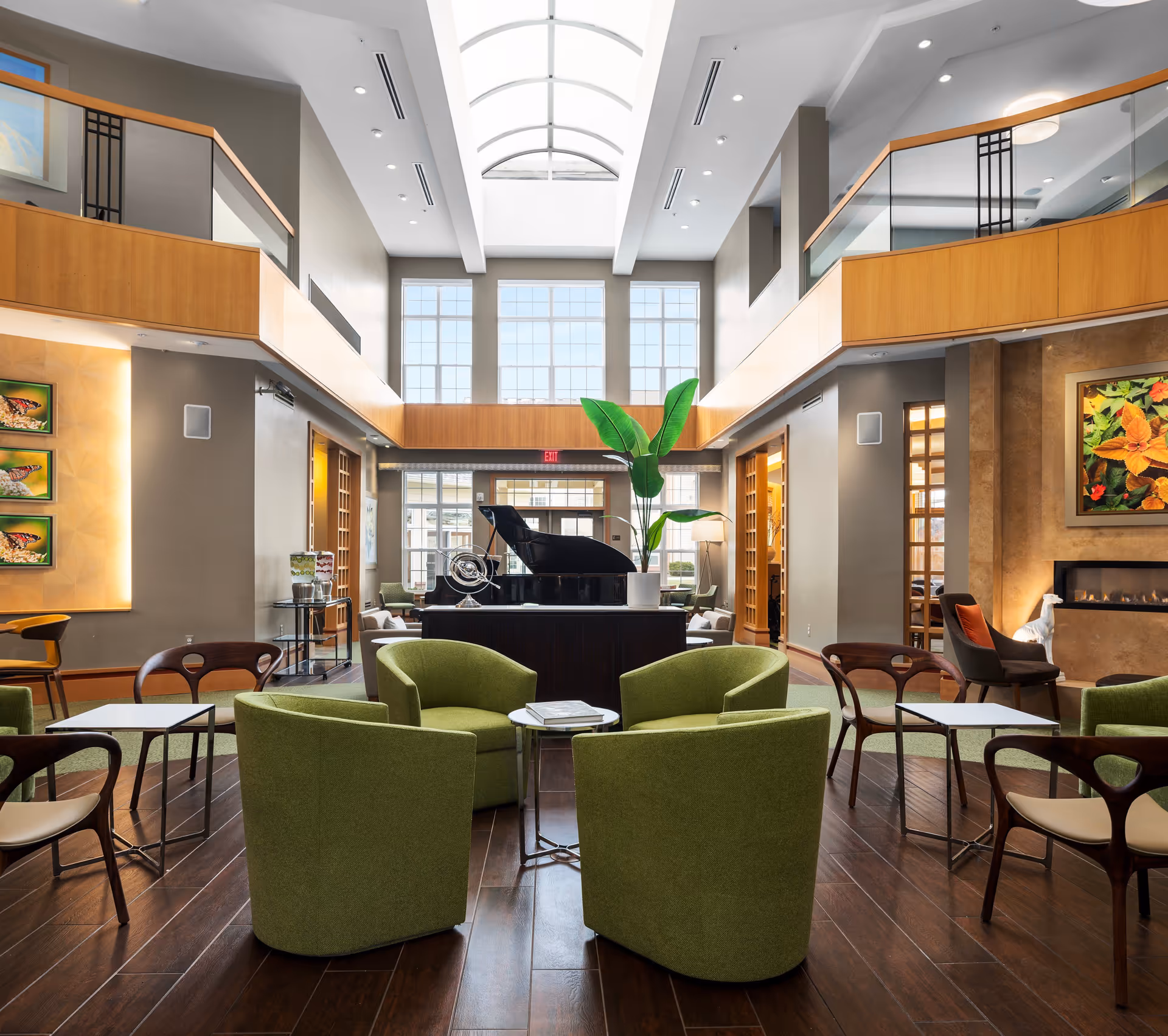 Bright two-story atrium lounge with green armchairs arranged around small tables, a grand piano, tall skylight, and a fireplace.