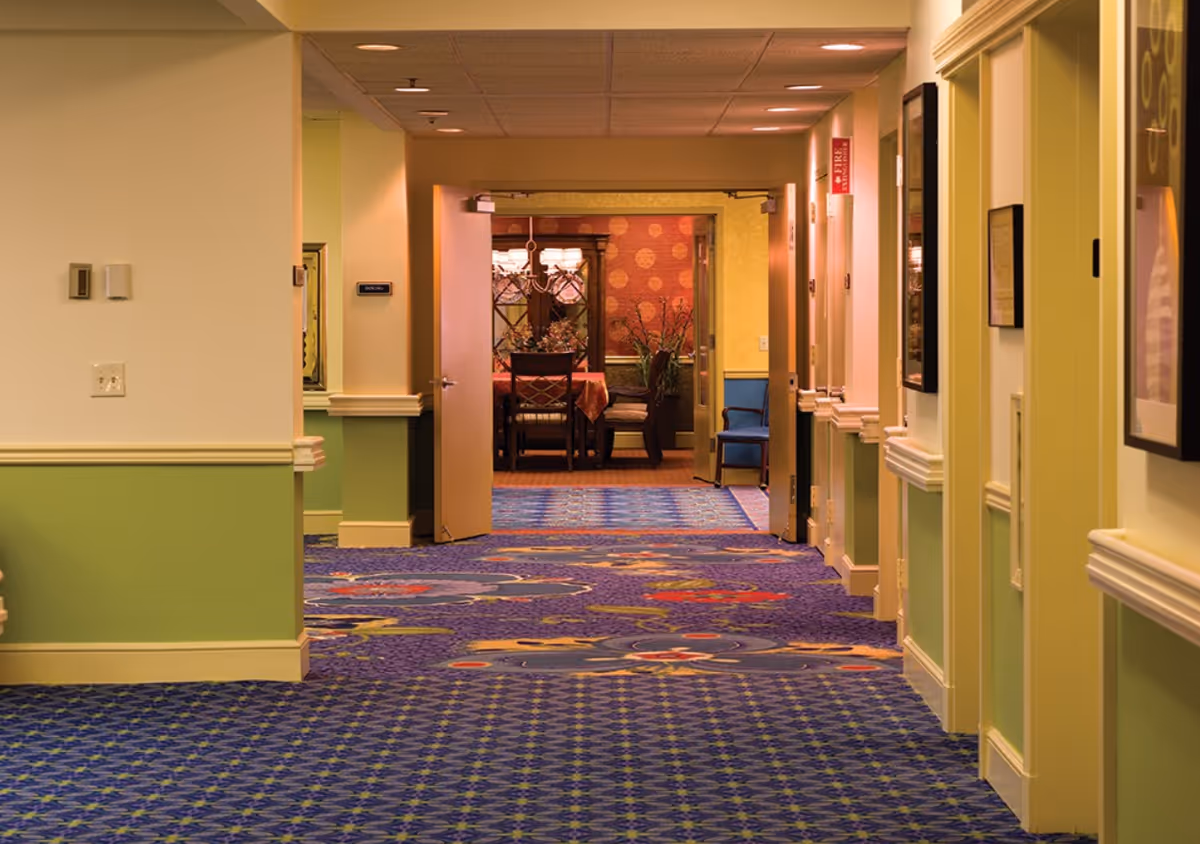 Carpeted interior hallway in an assisted living facility with green-and-cream walls and open double doors revealing a dining area with a table and chairs.