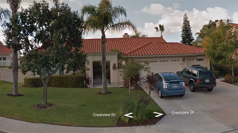 Single-story residential building with a red tile roof, a driveway with two parked cars, and a well-maintained front yard featuring palm trees and other greenery under a partly cloudy sky.