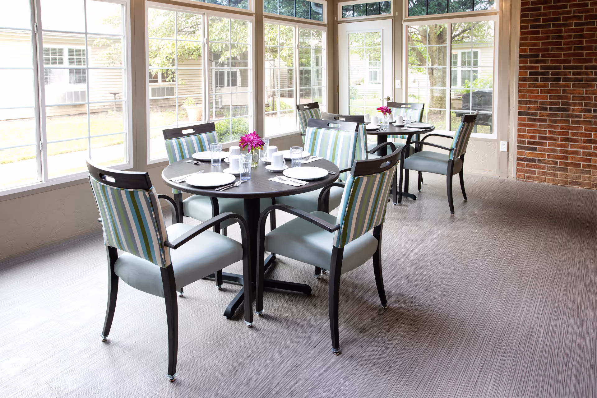 Sunlit dining area with round tables set for meals and striped upholstered chairs by large windows.