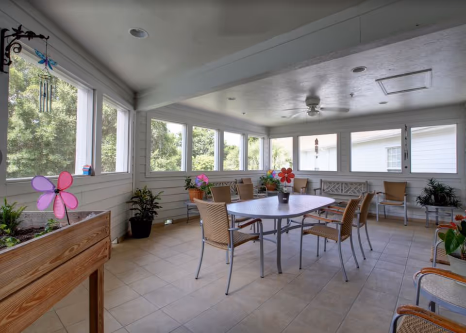 Enclosed sunroom with a central table surrounded by chairs, potted plants, and large windows overlooking greenery.