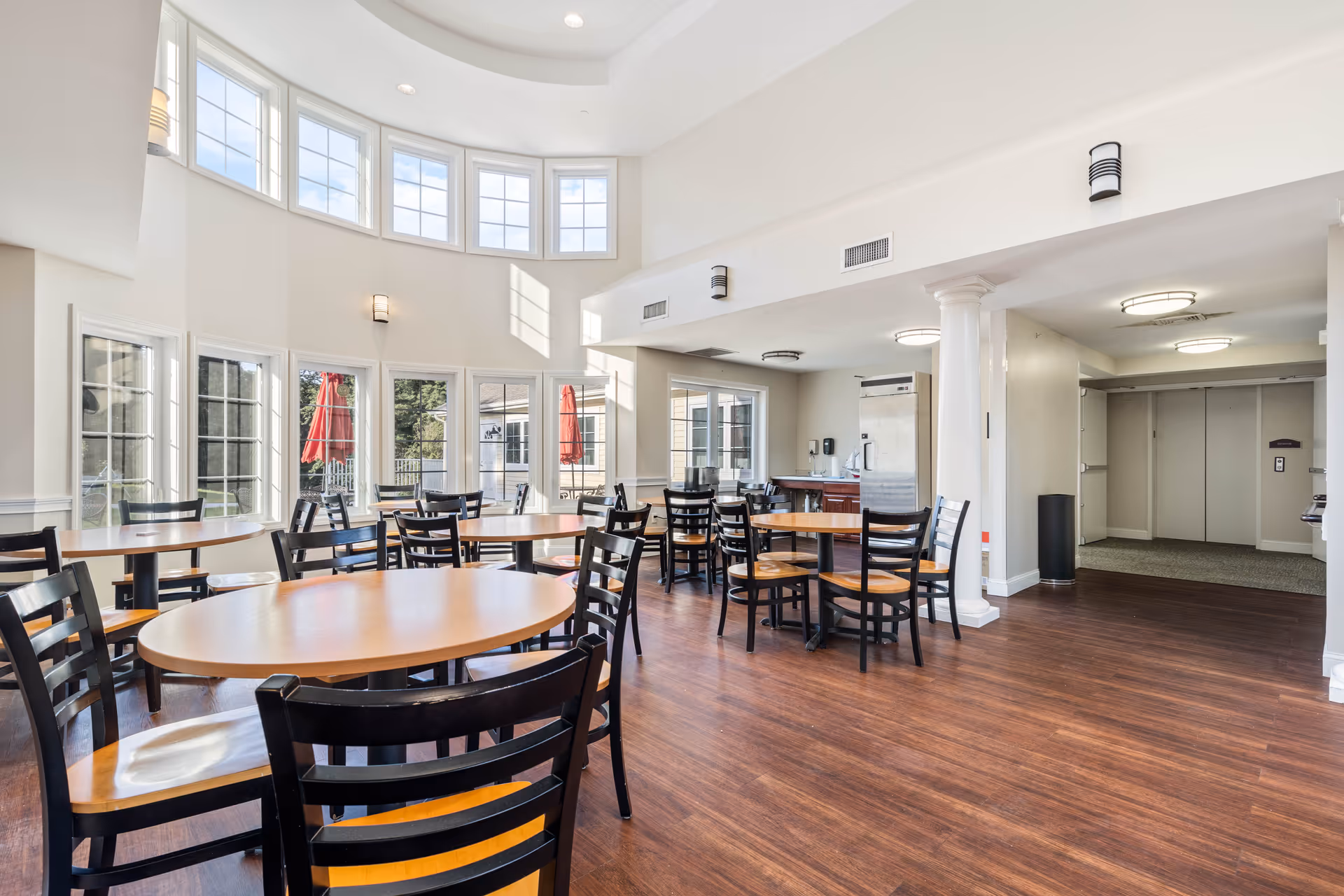 A bright dining room with multiple round wooden tables and black chairs arranged neatly on a wooden floor. Large windows and glass doors let in natural light, and red umbrellas are visible outside. The room has high ceilings with several light fixtures and white walls with decorative molding. There is an elevator and a stainless steel refrigerator in the background.
