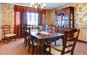 A traditional dining room with a wooden dining table surrounded by six chairs with cushioned seats. The room features floral wallpaper, a wooden china cabinet with glass doors, a chandelier hanging from the ceiling, and a window with sheer and dark red curtains.