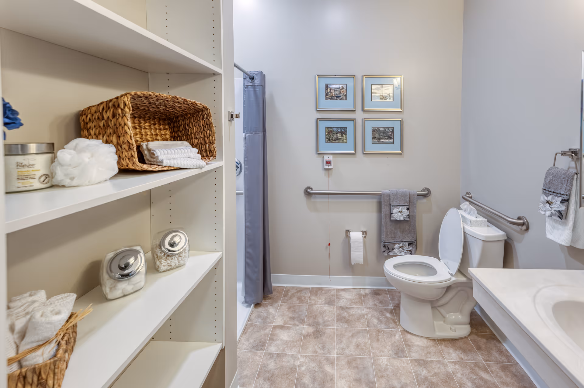 A clean and accessible bathroom with a toilet, grab bars, a shower with a gray curtain, and white shelving stocked with towels and toiletries. The walls are decorated with four framed pictures, and the floor has beige tiles.