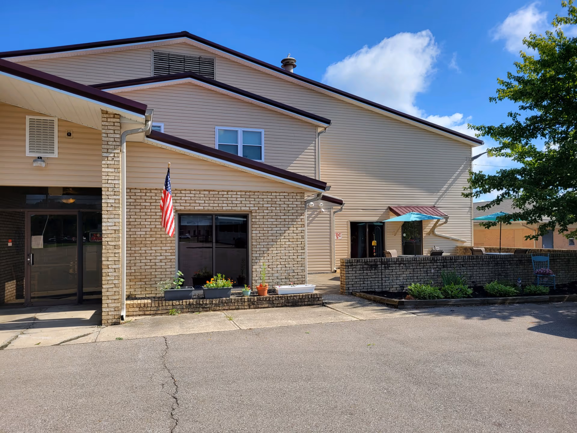 Exterior view of a beige two-story assisted living facility building with a brick lower facade. There is an American flag mounted on the wall near a window with flower pots on the ledge. To the right, there is a small patio area with blue umbrellas and outdoor seating. The sky is clear with some clouds, and a tree is visible on the right side.