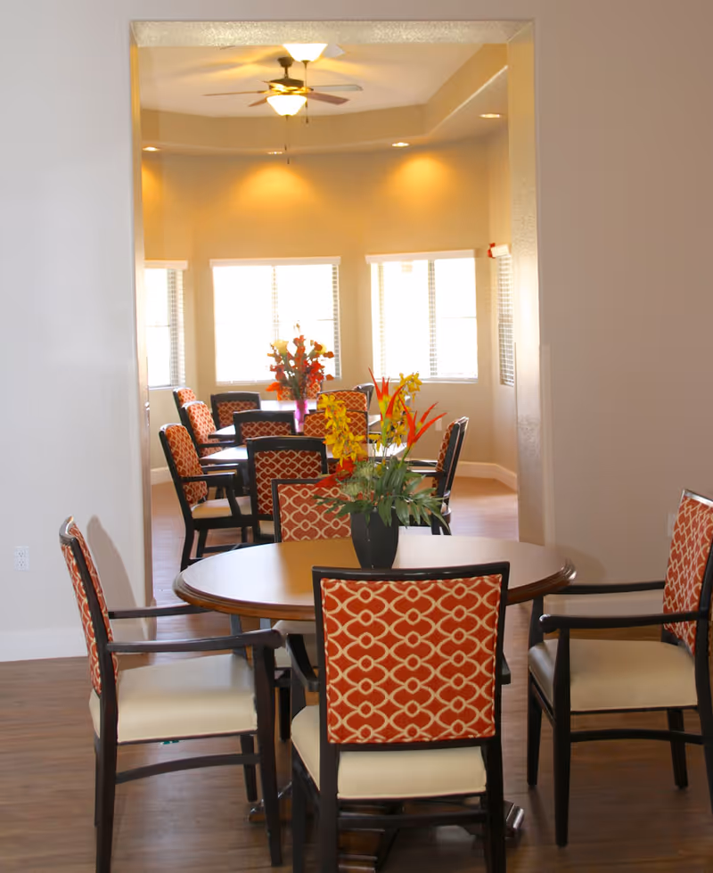 Communal dining room with round tables, orange-patterned chairs, and floral centerpieces under recessed lighting.