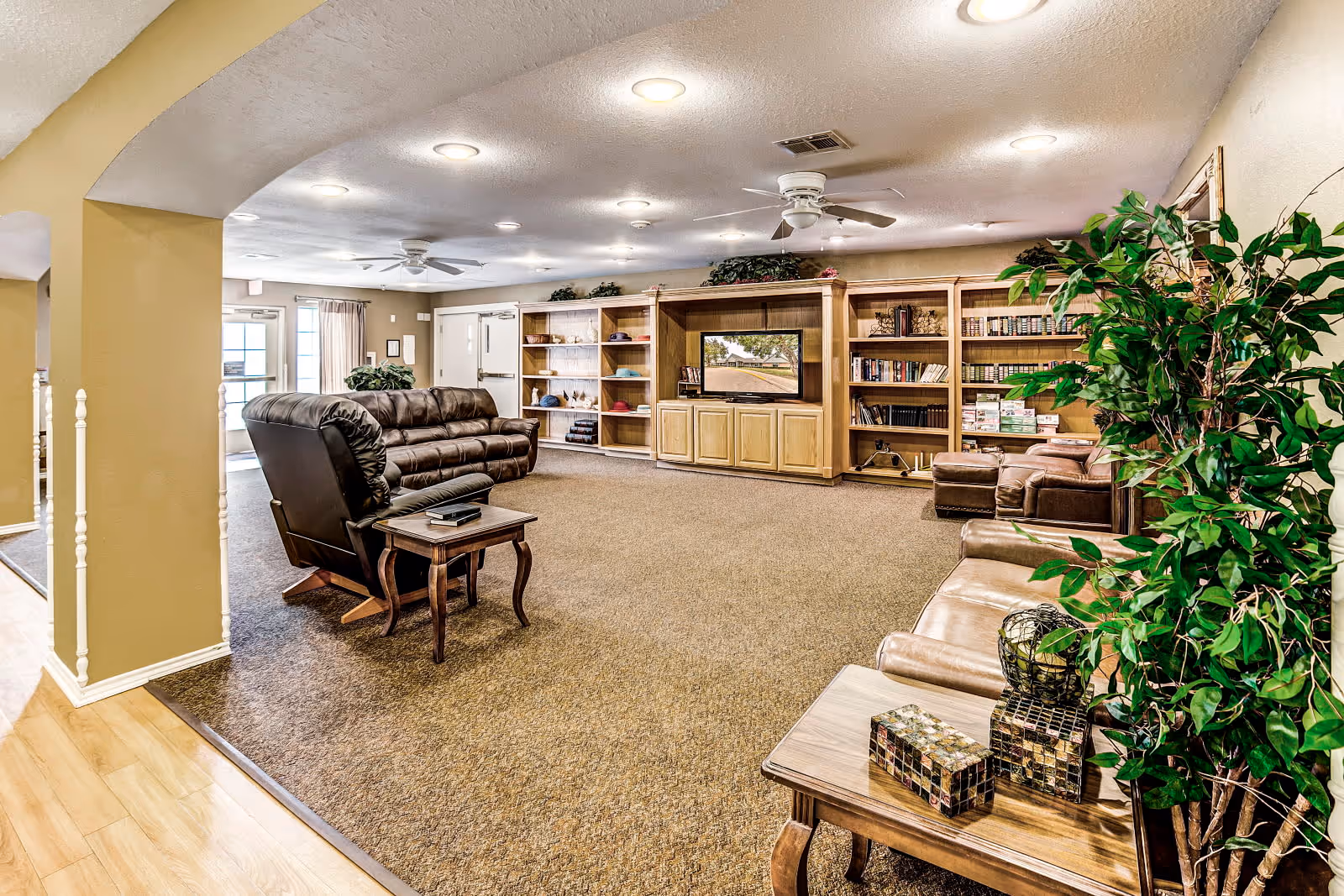 A spacious living room area in an assisted living facility featuring brown leather sofas and armchairs, wooden side tables, a large built-in wooden entertainment center with a TV and bookshelves, ceiling fans, recessed lighting, and a large green plant in the foreground.