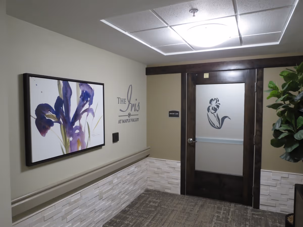 Interior hallway of a senior living facility with a door labeled Memory Care featuring a frosted glass panel with a flower design. On the left wall, there is a framed painting of purple irises and the text 'The Iris at Maple Valley'. A green plant is partially visible on the right side.