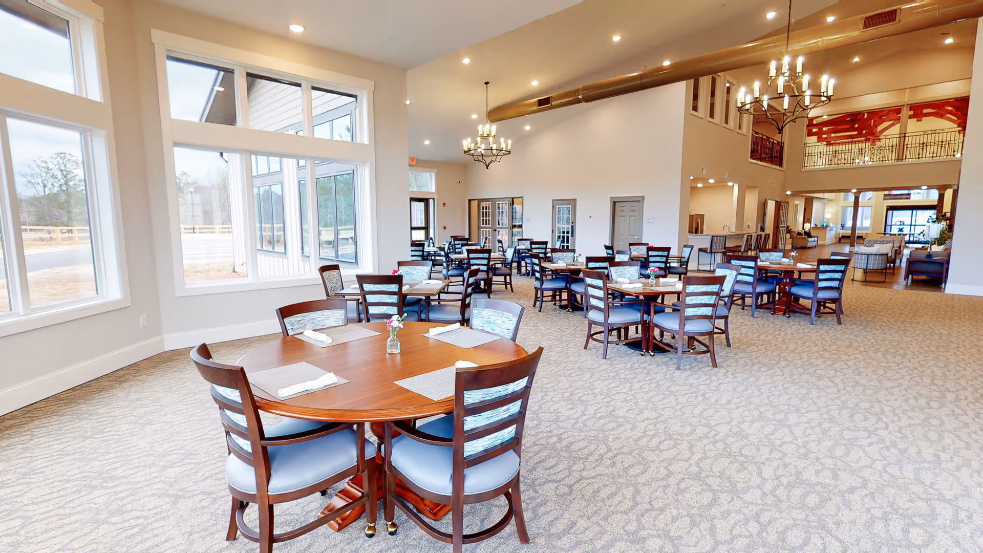 A spacious senior living dining room with multiple wooden tables and chairs arranged neatly. Each table has placemats, napkins, and small flower vases. Large windows on the left side let in natural light, and chandeliers hang from the ceiling. The room has a carpeted floor and an open layout leading to other common areas.