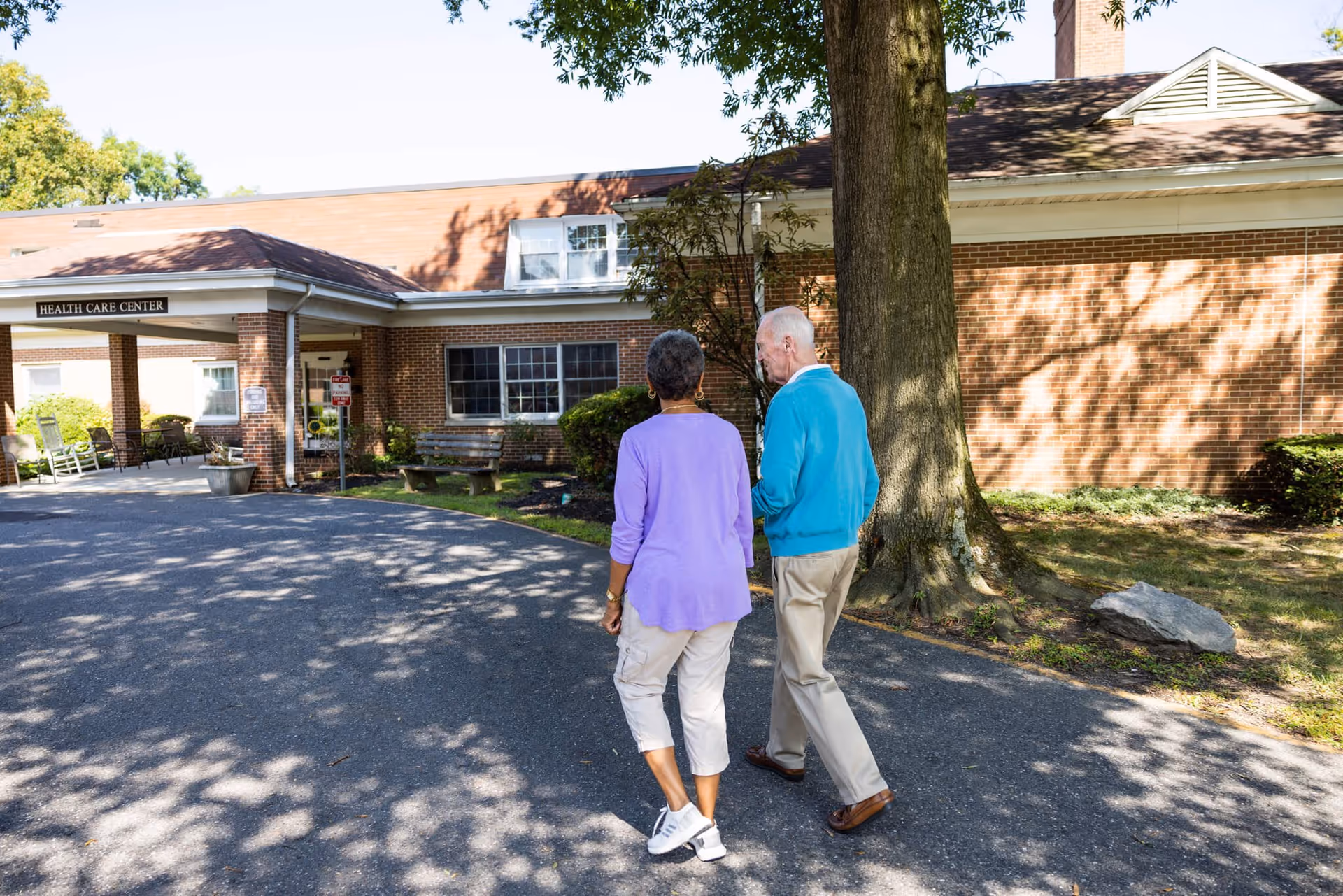 An elderly man and woman walking together on a paved path outside a brick building with a sign that reads 'Health Care Center'. The area is shaded by trees and there are benches and rocking chairs near the entrance.