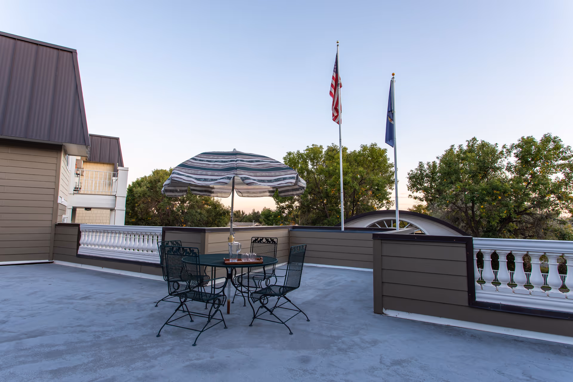 Rooftop patio with a round table and metal chairs under a striped umbrella, two flagpoles, and trees in the background.