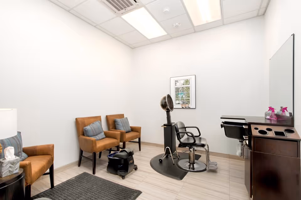 A small salon room with a black salon chair and hair dryer, two brown armchairs with blue cushions, a small table with a tissue box and lamp, a dark wooden cabinet with spray bottles, and a framed picture on the white wall.