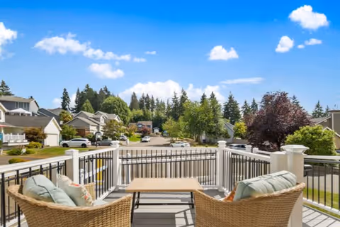 View from a balcony with two wicker chairs with cushions and a small wooden table between them, overlooking a suburban neighborhood street lined with houses, trees, and parked cars under a bright blue sky with scattered clouds.
