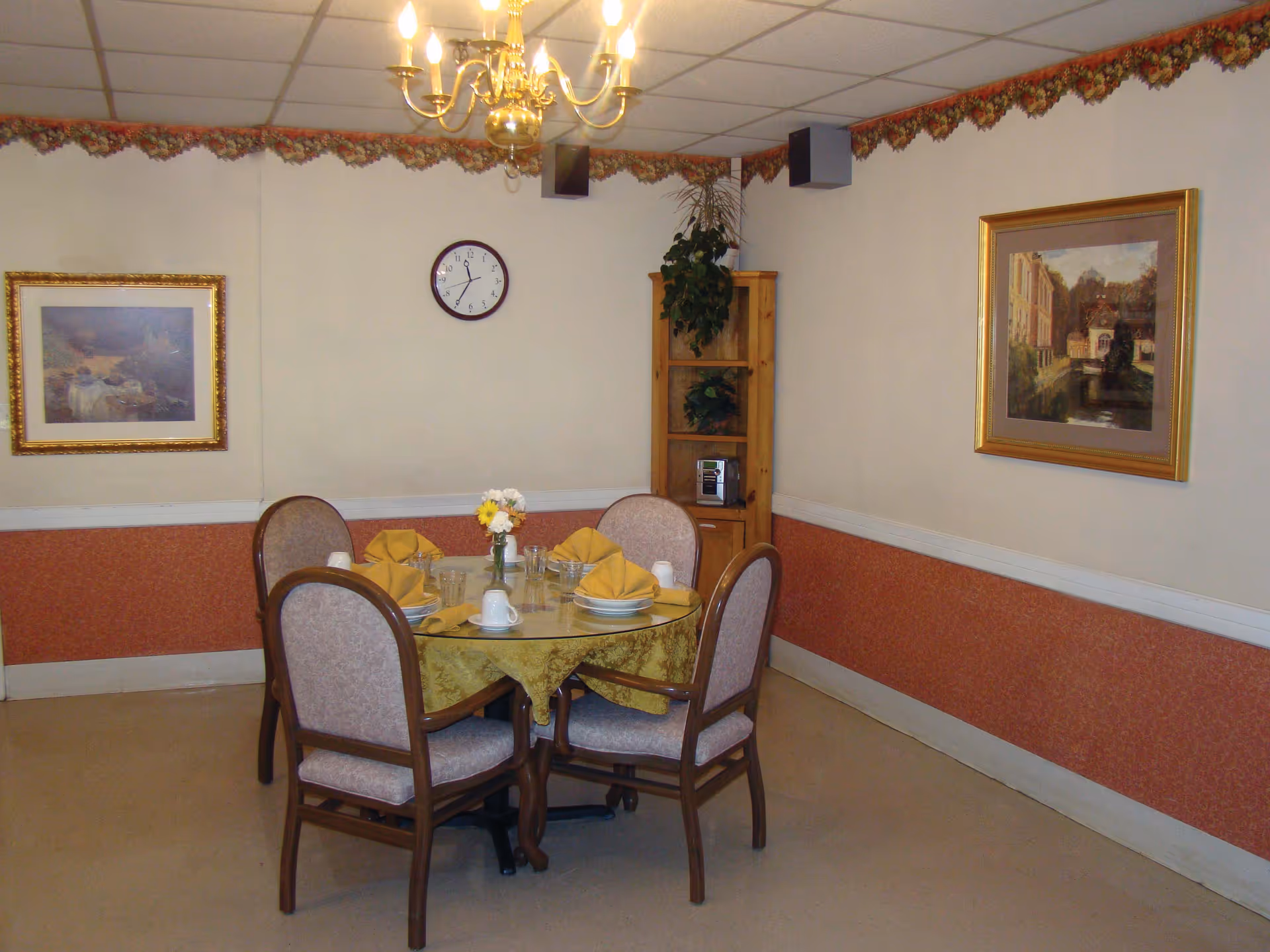 Round dining table set for four in a small dining room with a chandelier, wall clock, framed paintings, and a wooden shelf.