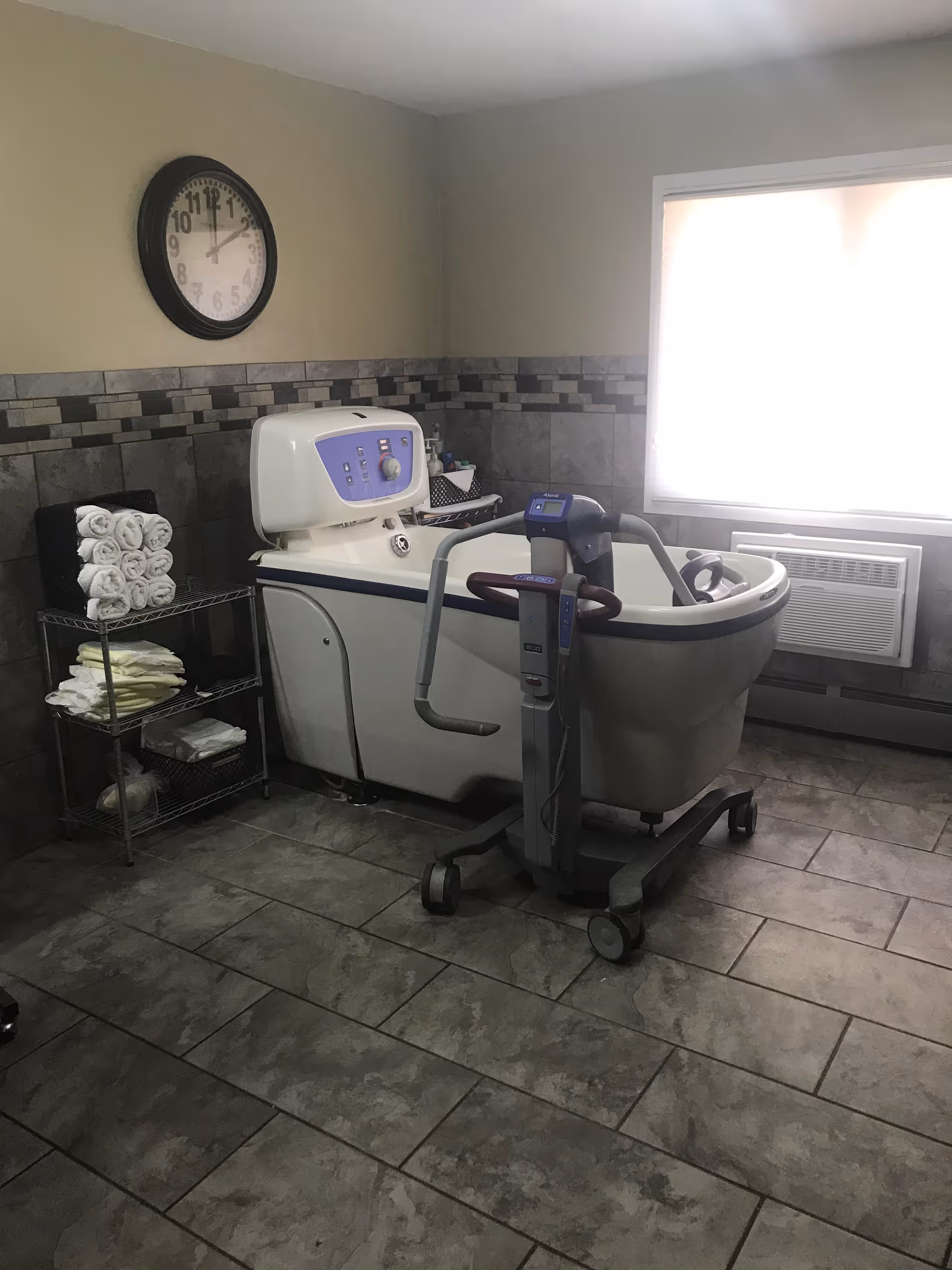 A tiled bathing room with a medical bathtub and patient lift, shelving of rolled towels, a wall clock, and a bright window.