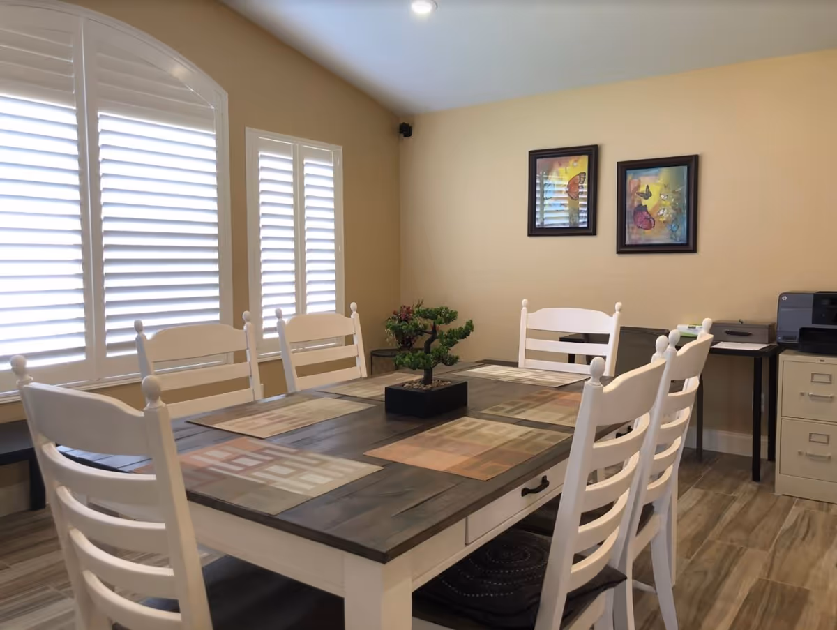 Bright dining room with a rectangular wooden table surrounded by white chairs, plantation shutters and framed artwork on a beige wall.