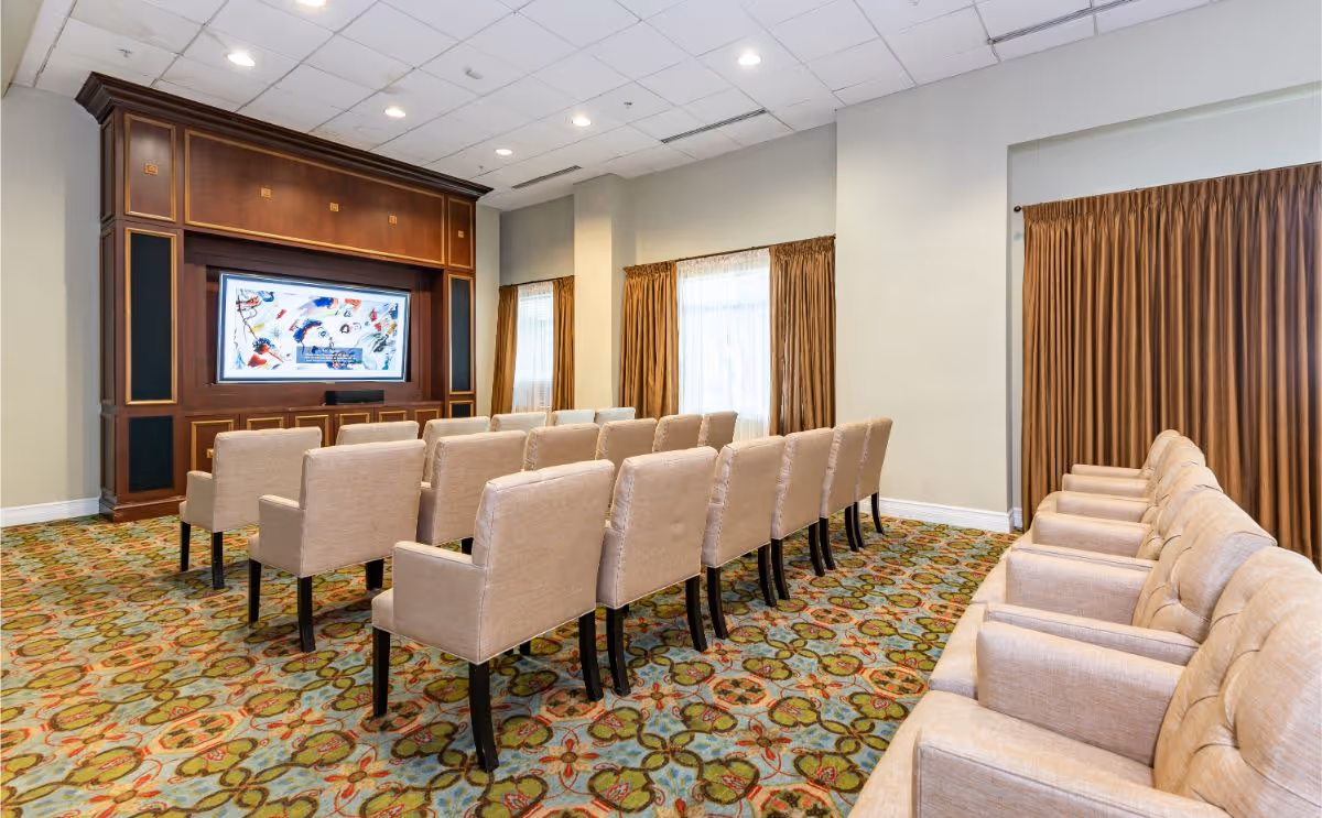 A carpeted community media room with rows of upholstered chairs facing a large screen set in wood paneling.