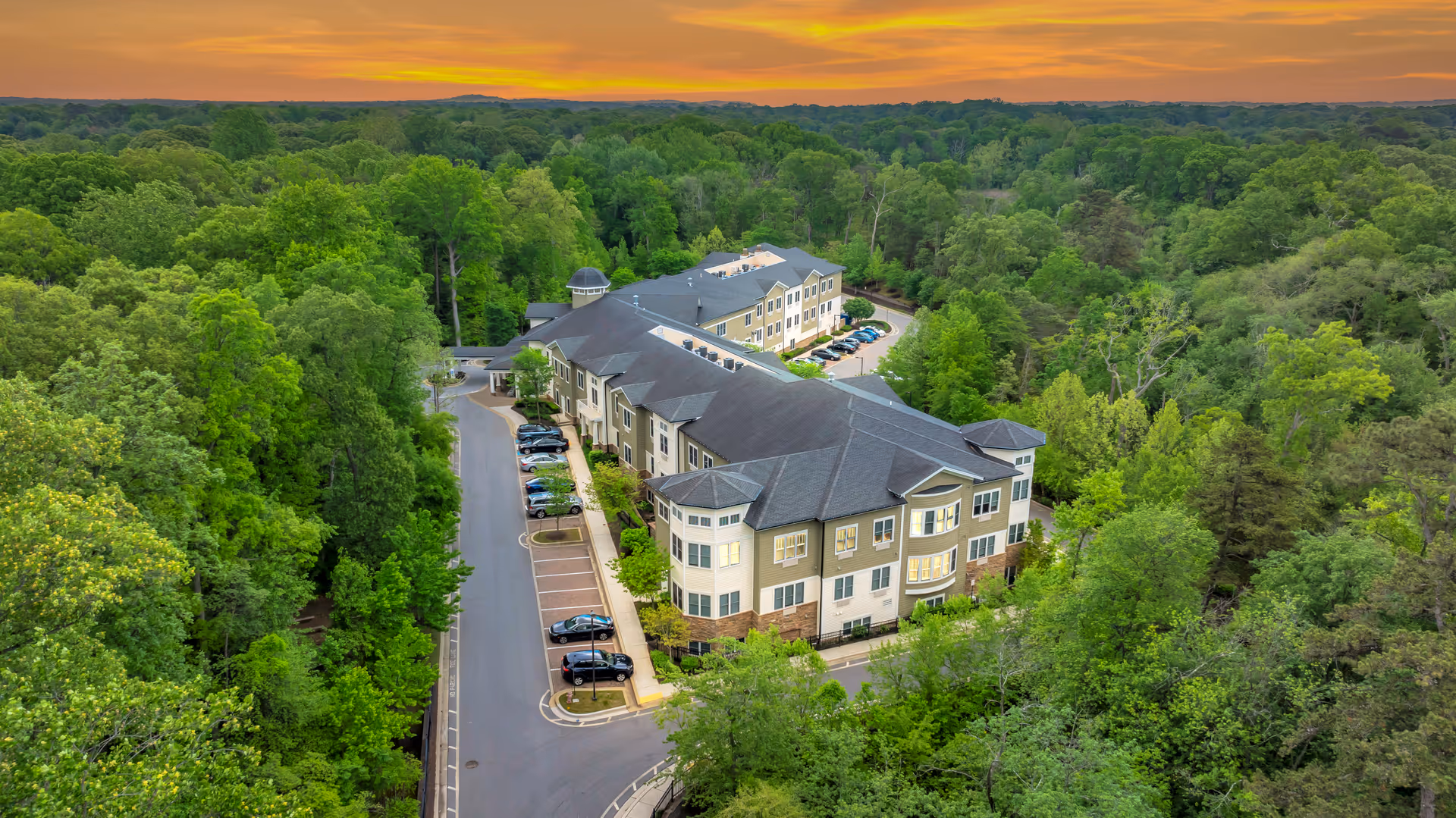 Aerial view of Spring Arbor of Severna Park, a multi-story senior living facility surrounded by dense green trees under a vibrant orange sunset sky. The building has a gray roof and beige exterior with multiple windows, and there is a parking area with several cars parked along the driveway.
