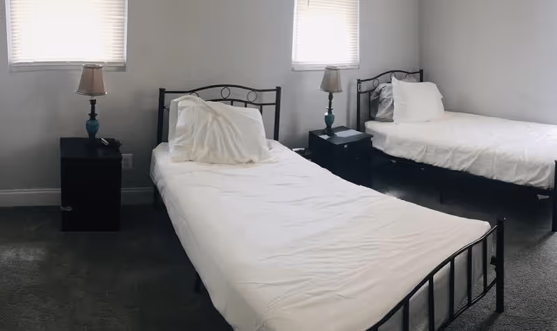 A simple bedroom with two single metal-frame beds dressed in white linens, two nightstands with lamps, and two windows.