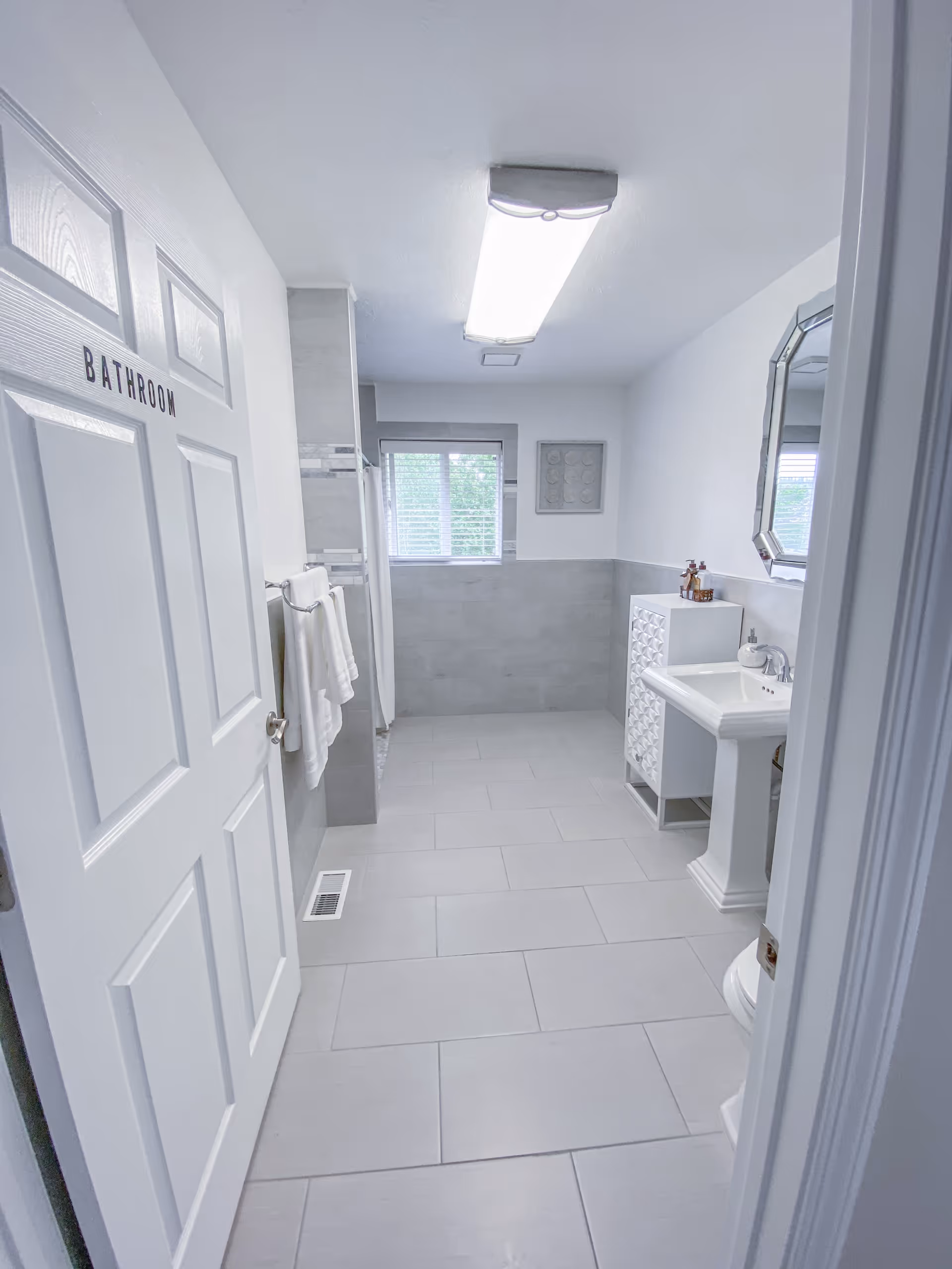 Bright white and gray bathroom viewed from the doorway, featuring a pedestal sink, mirror, towel rack, and a door labeled 'BATHROOM'.