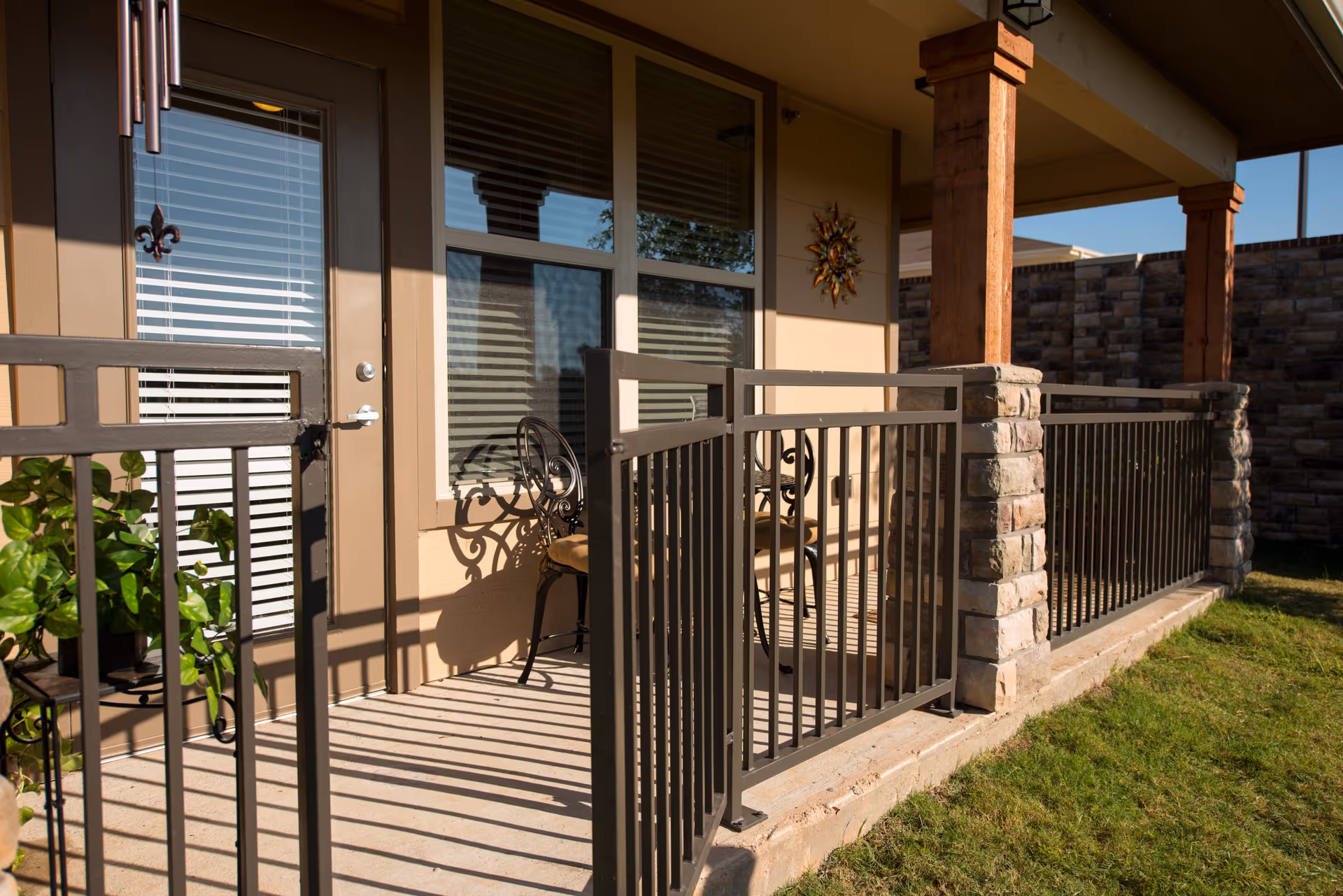 A small gated patio with metal railings and stone columns, featuring bistro chairs, a door and windows opening onto a lawn.