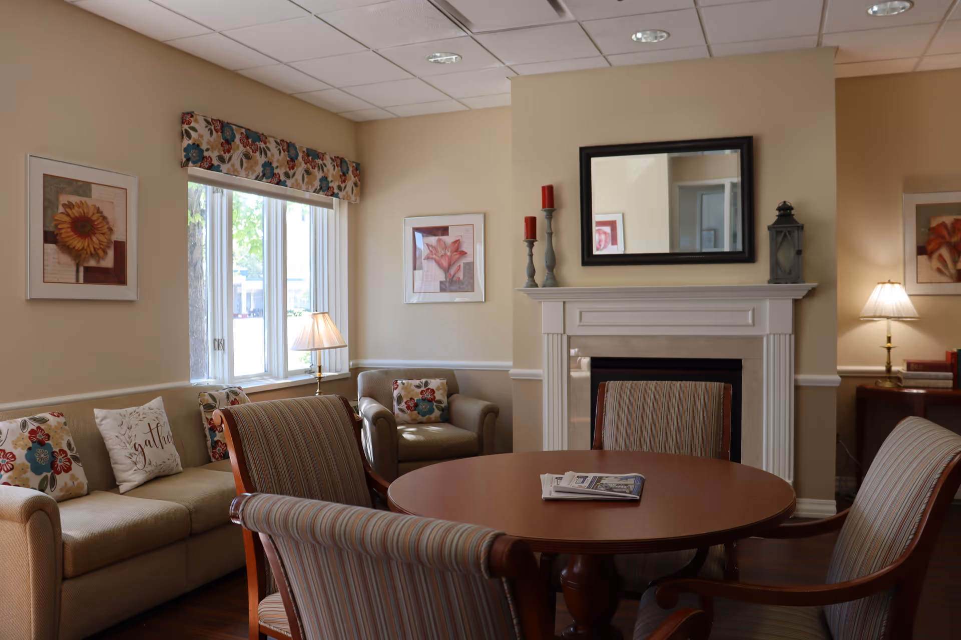 A cozy living room area with a round wooden table surrounded by four striped upholstered chairs. Behind the table is a beige sofa with floral and decorative pillows, a matching armchair, and a window with floral valance. The room features a white fireplace with a black-framed mirror above it, red candles on candle holders, and a lantern. There are framed floral artworks on the walls and two table lamps providing warm lighting.