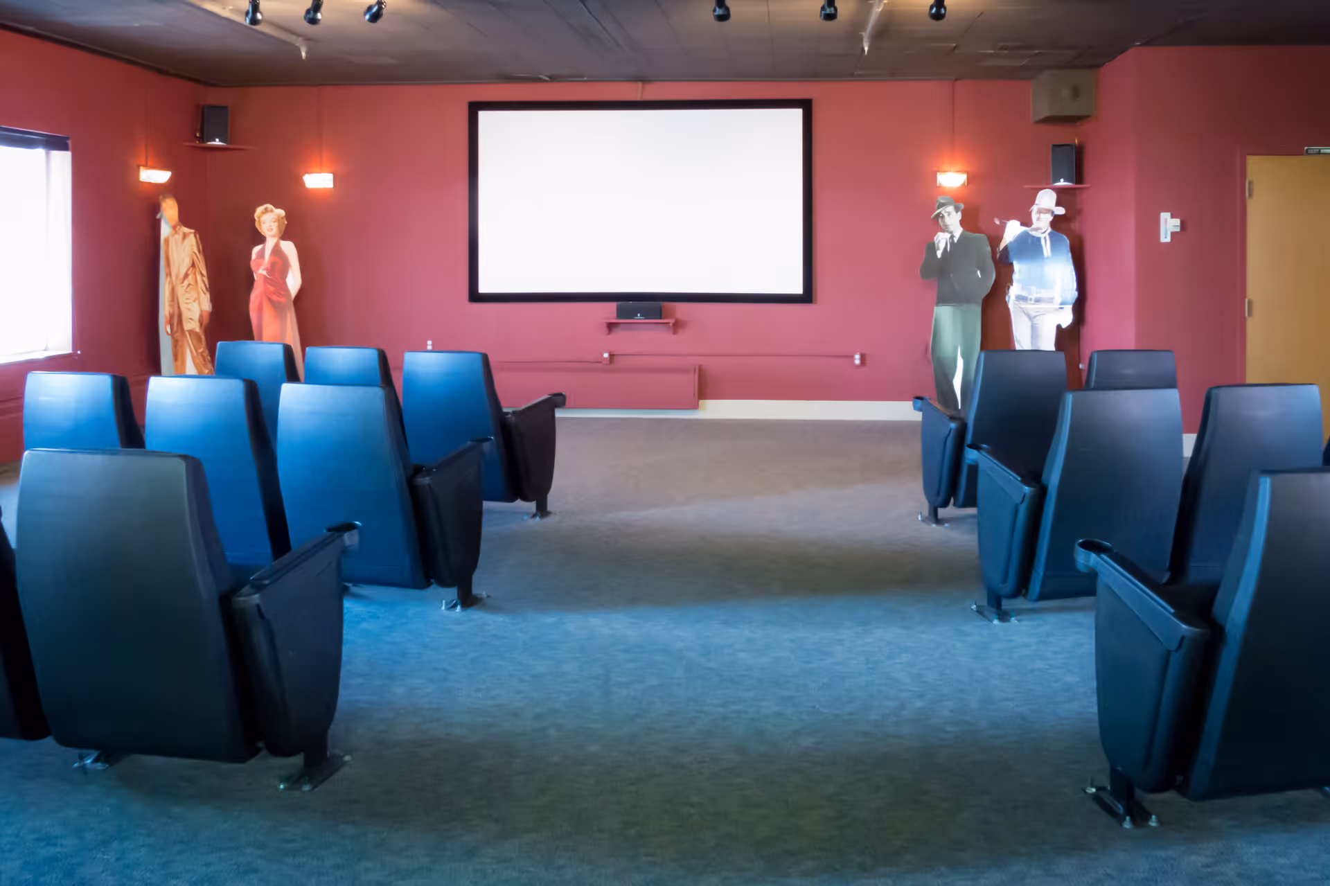 A small movie theater room with rows of black cushioned seats facing a large blank white screen on a red wall. There are four life-sized cardboard cutouts of vintage movie characters positioned near the walls. The room has a carpeted floor and ceiling lights.
