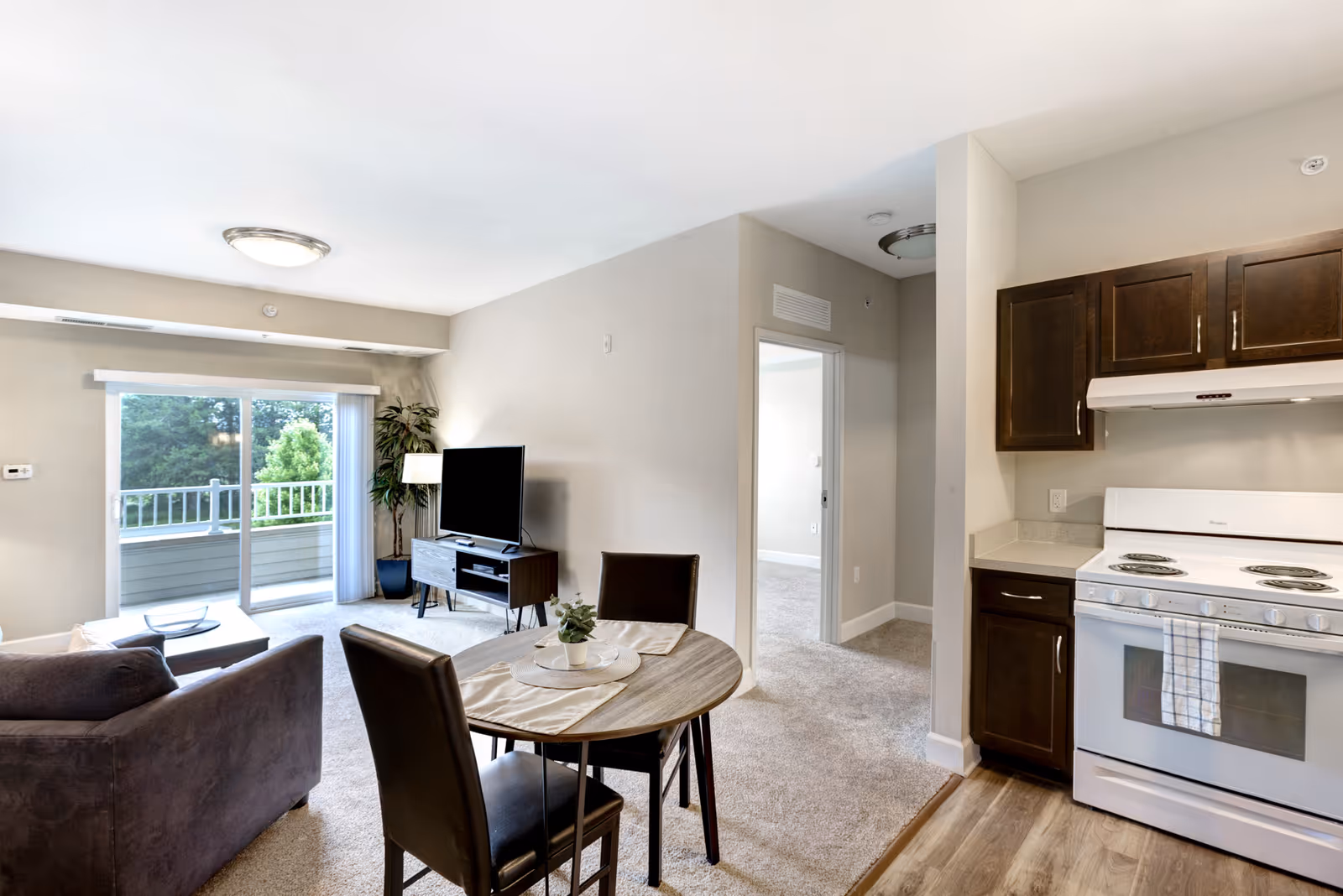 Open-plan living area with a sofa, TV on a stand, a small dining table, and a kitchen with a white stove, plus sliding glass doors to a balcony.