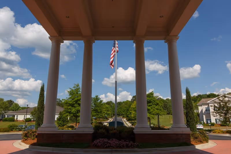 View from under a large covered entrance with four white columns, looking out towards an American flag on a flagpole, surrounded by trees, shrubs, and buildings under a partly cloudy blue sky.