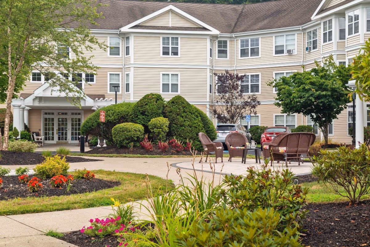 Outdoor courtyard area of a senior living facility with a paved walkway, landscaped garden beds with colorful flowers and shrubs, and a seating area with wicker chairs and a table. The beige multi-story building with many windows surrounds the courtyard.